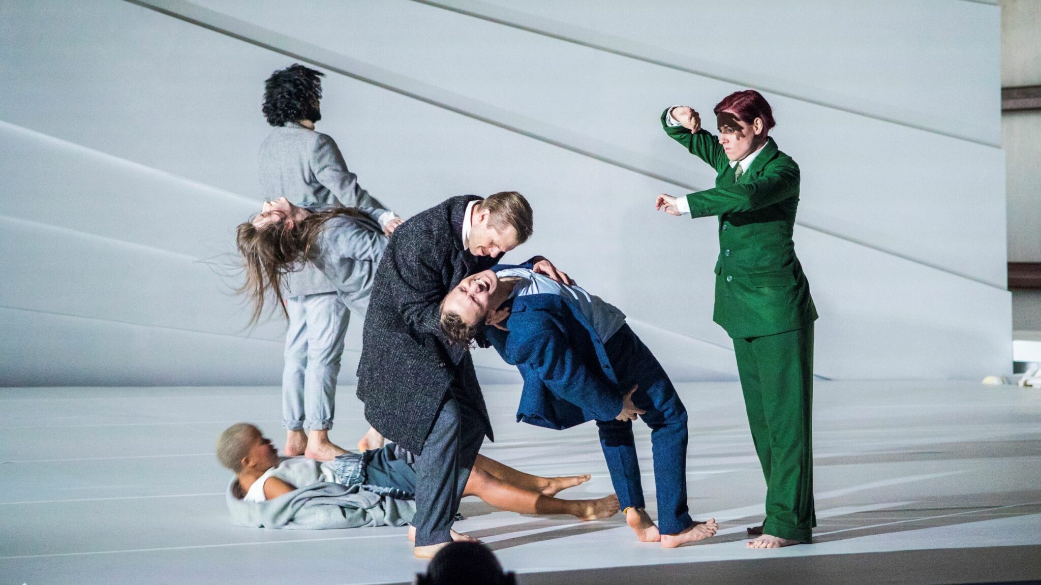 six people on a white stage. A man is falling and another man is catching him.