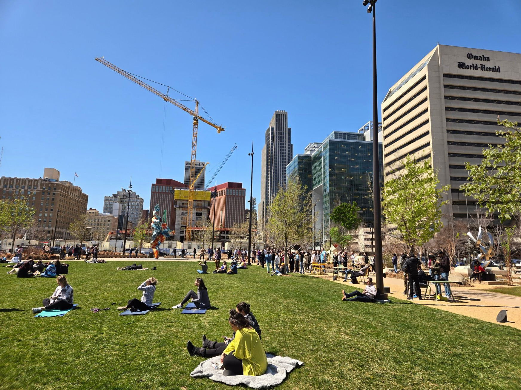 An image of a downtown park with trees in the spring. People are gathered and sitting on blankets.